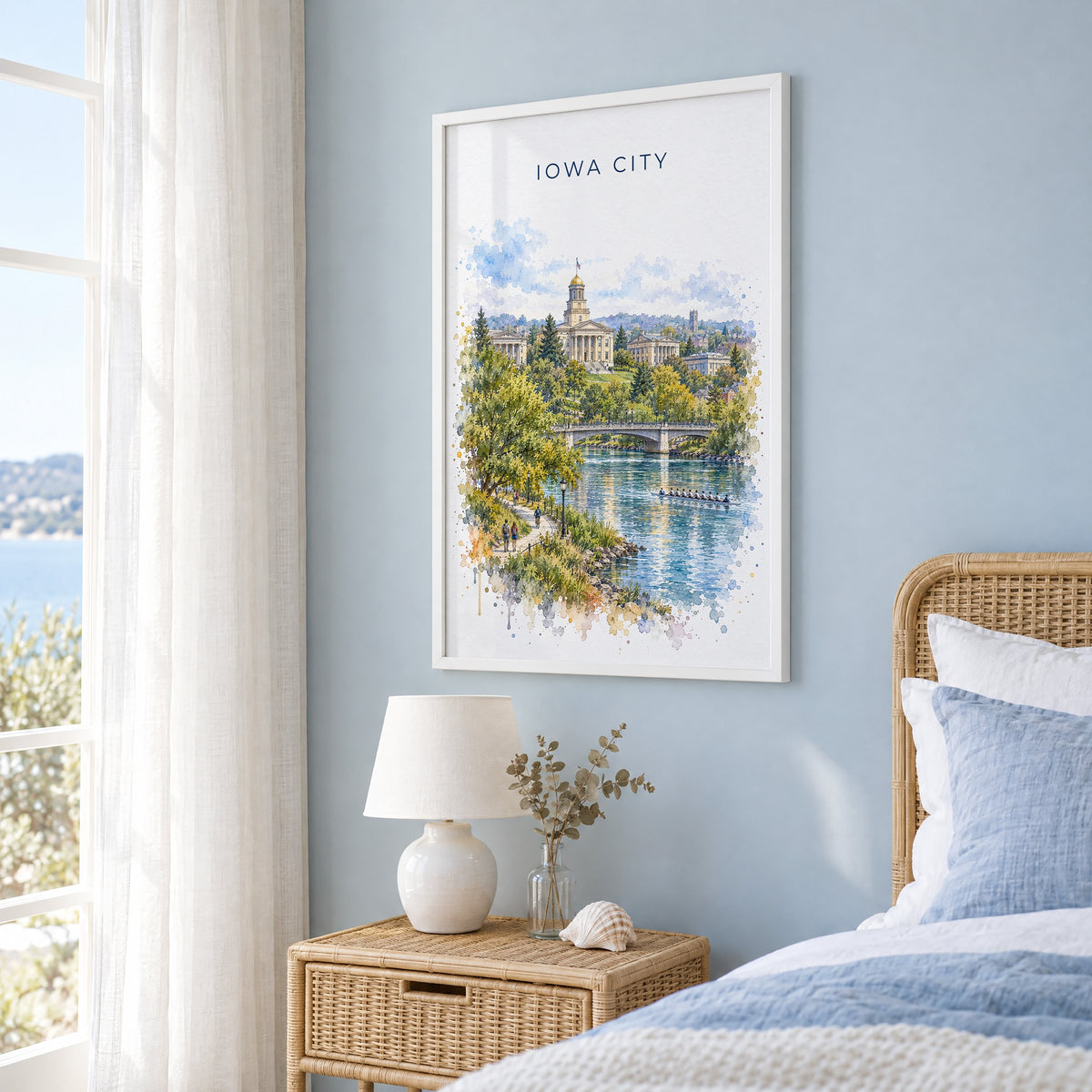Iowa City watercolor print in a white frame on a pale blue wall beside a rattan bed in a coastal bedroom