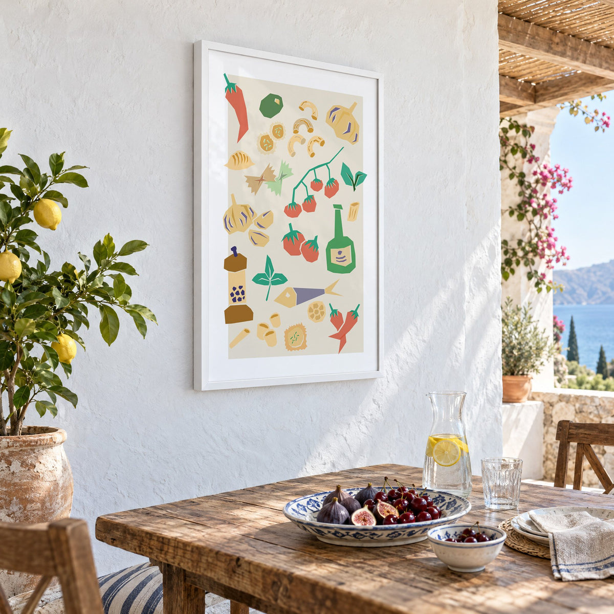 Italian ingredients cut-paper pattern print in a white frame on a whitewashed terrace wall above a lunch table with figs and cherries, seen at an angle