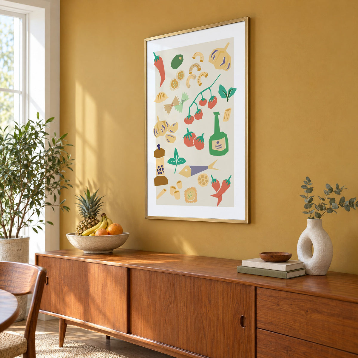 Italian ingredients cut-paper pattern print in a brass frame on a mustard wall above a teak sideboard with a fruit bowl, seen at an angle