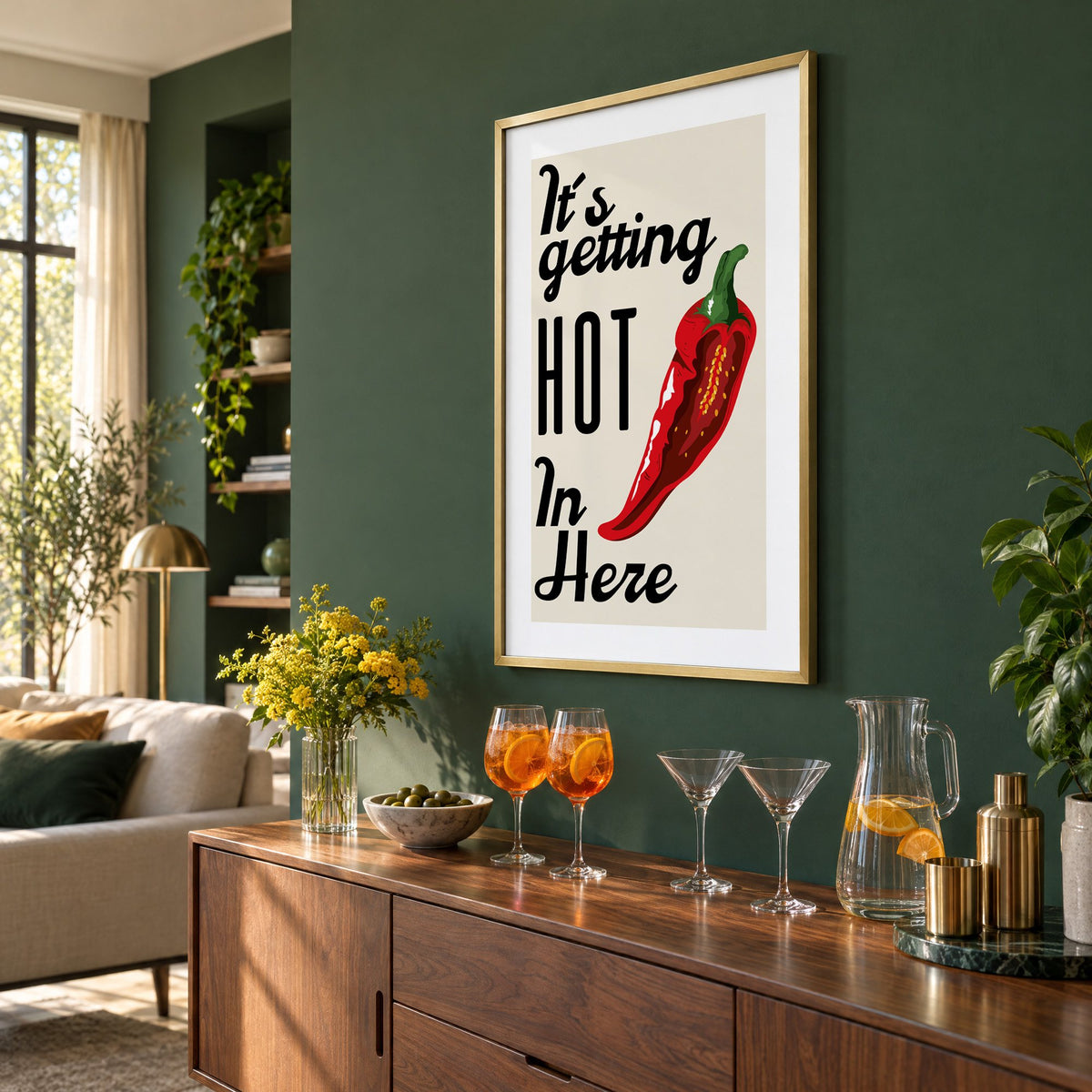 It's getting HOT In Here split chili print in a brass frame on a bottle green wall above a sideboard with spritz and martini glasses, seen at an angle