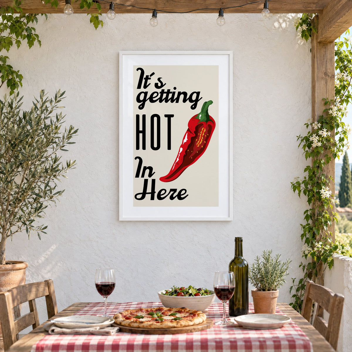 It's getting HOT In Here split chili print in a white frame on a stucco wall above a checked-cloth table with pizza and red wine