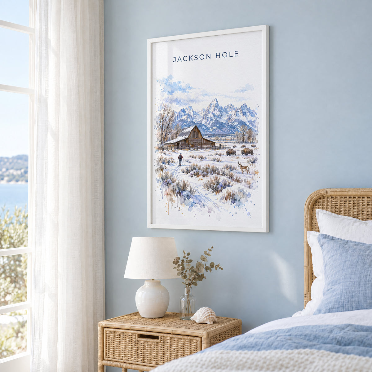 Jackson Hole watercolor print in a white frame on a pale blue wall beside a rattan bed in a coastal bedroom