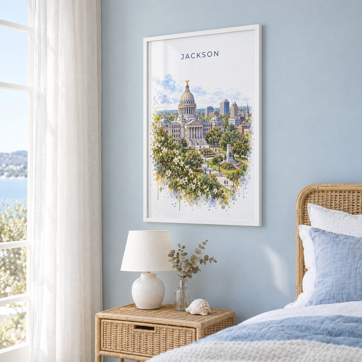 Jackson, Mississippi watercolor print in a white frame on a pale blue wall beside a rattan bed in a coastal bedroom