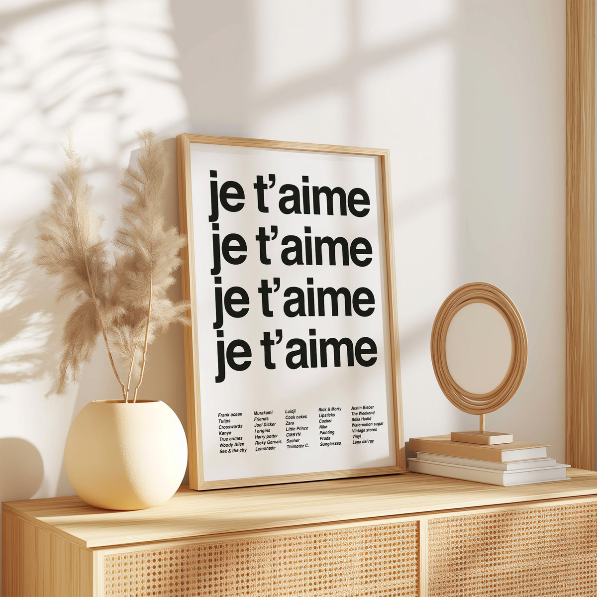 Repeated je t'aime black typography on white print in an oak frame on a rattan sideboard beside pampas grass