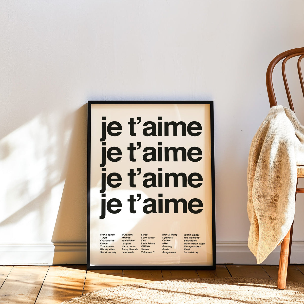 Repeated je t'aime black typography on white print in a black frame leaning on a wood floor beside a bentwood chair