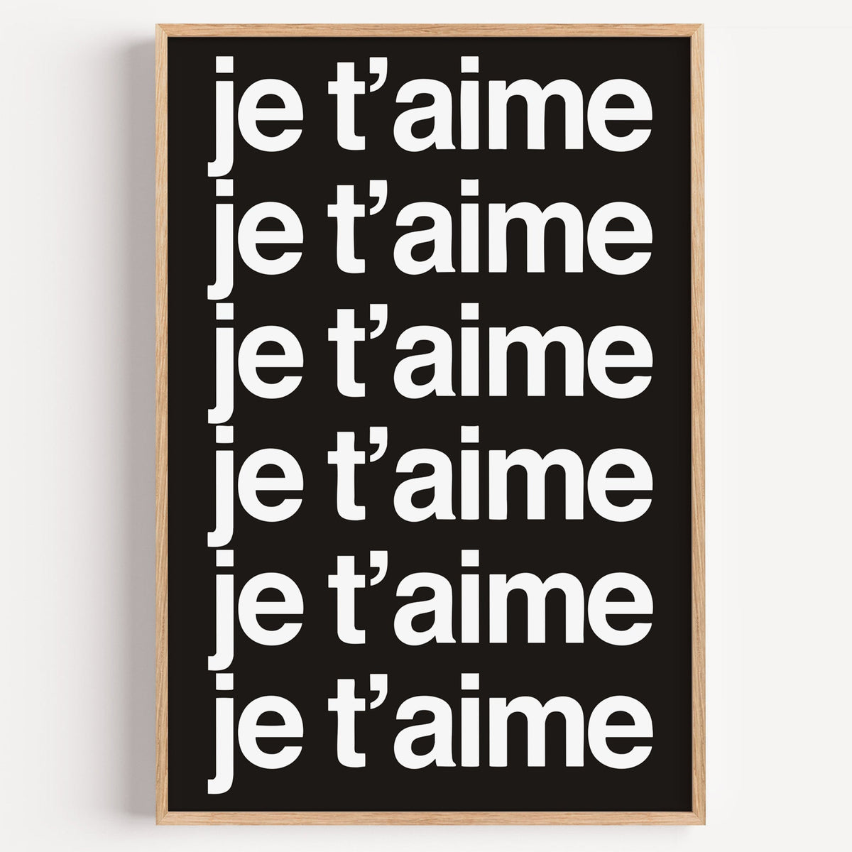 Je t'aime repeated six times in white type print in a light oak frame on a plain white wall