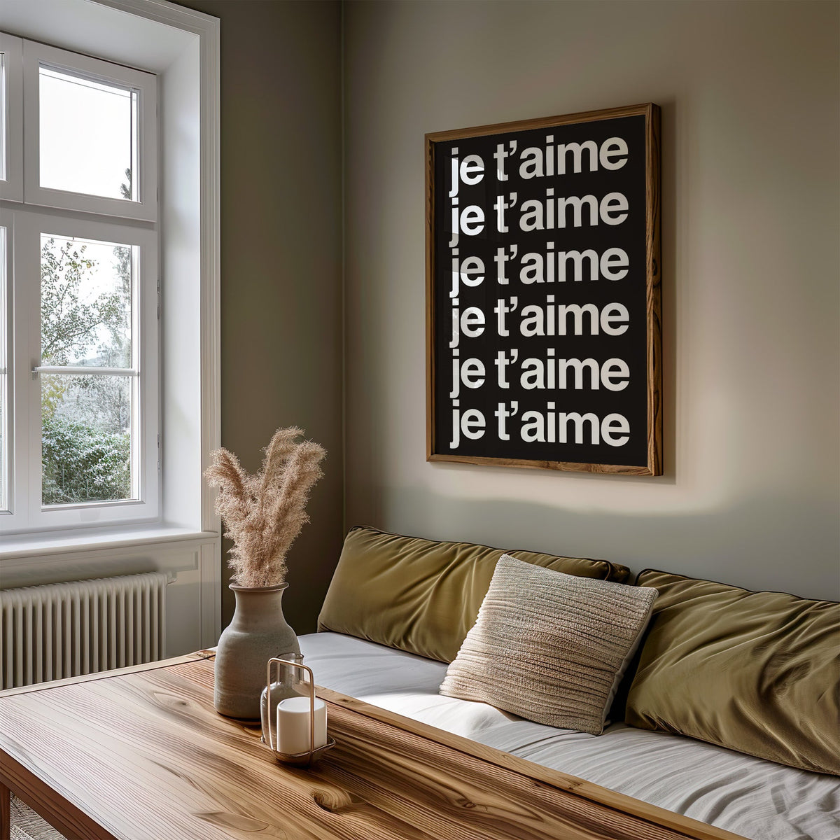 Je t'aime repeated six times in white type print in a walnut frame above an olive daybed by a window