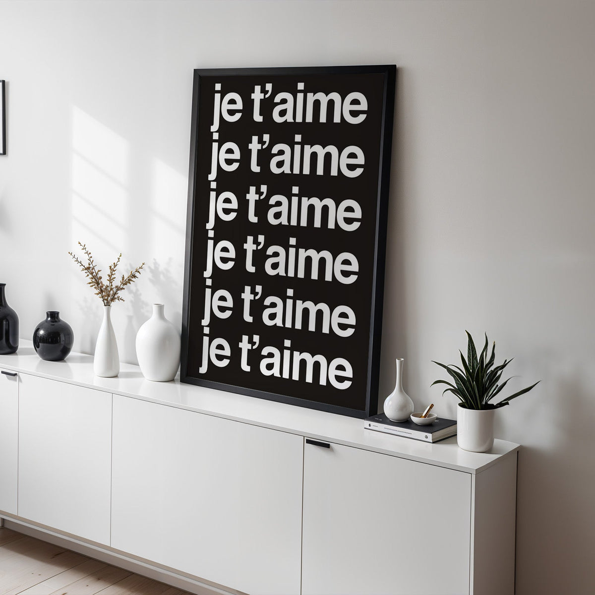 Je t'aime repeated six times in white type print in a black frame on a white sideboard with black and white vases