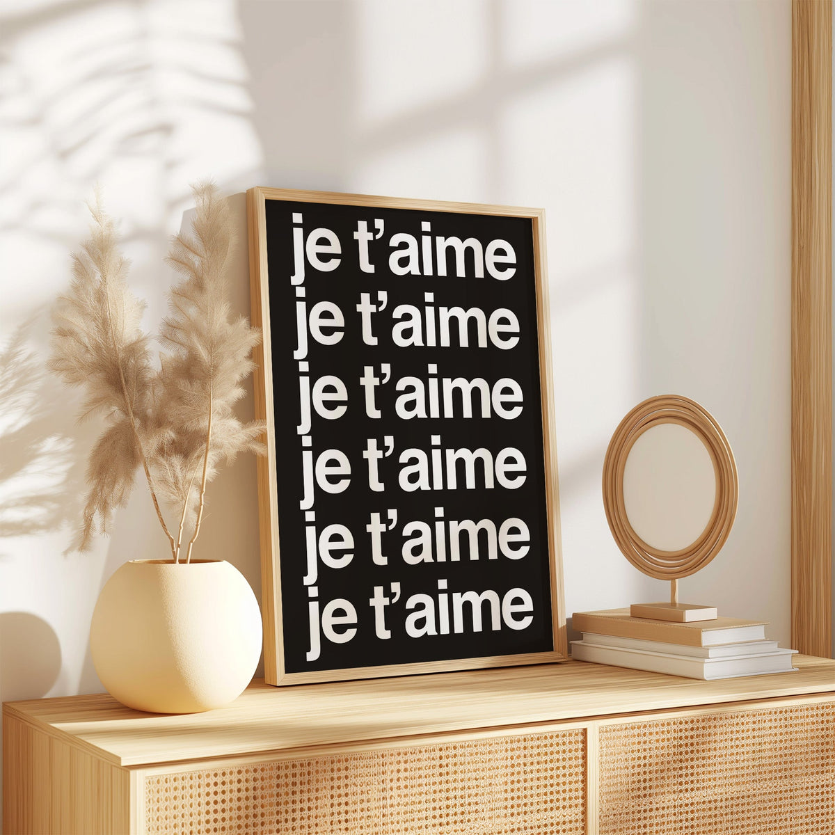 Je t'aime repeated six times in white type print in an oak frame on a rattan sideboard beside pampas grass