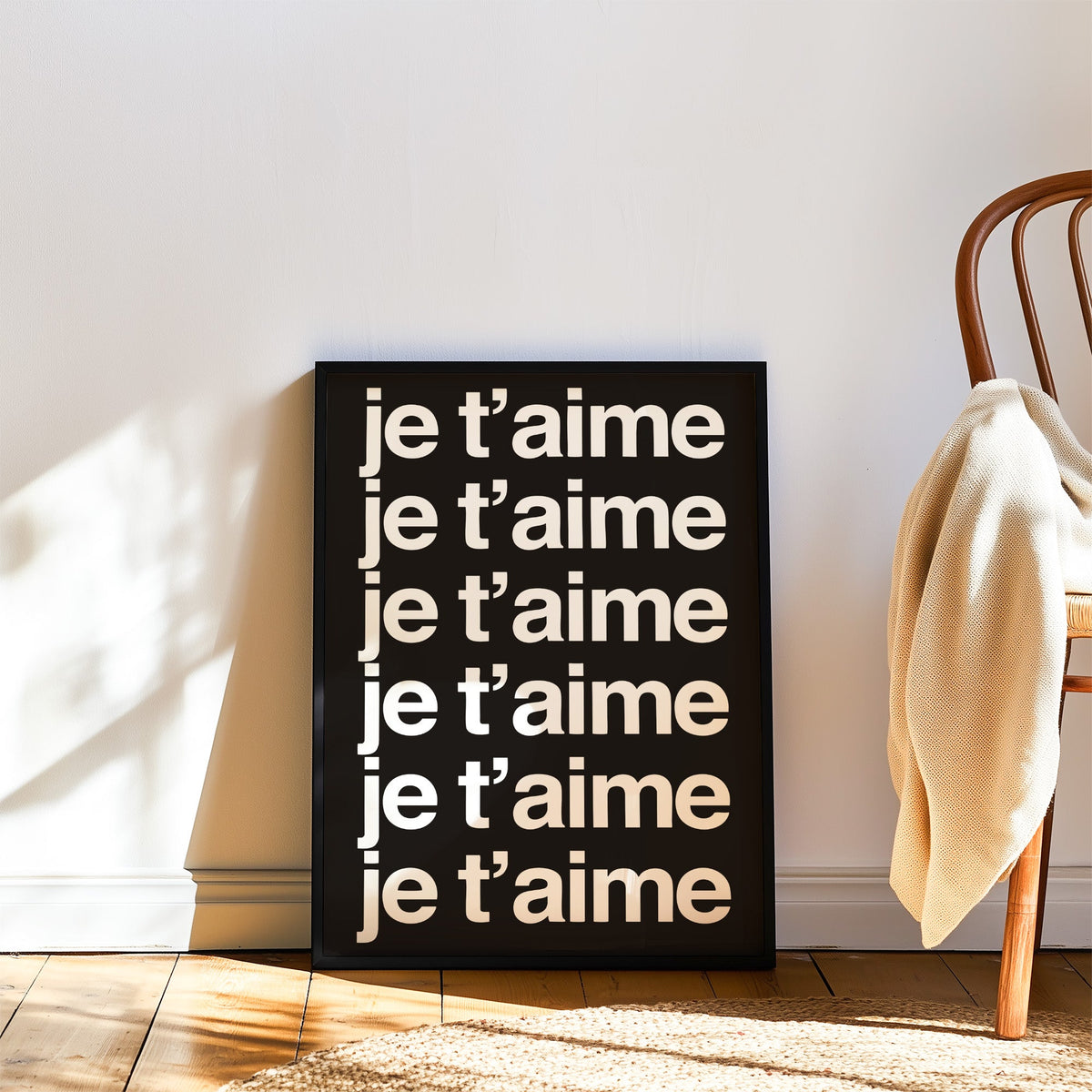 Je t'aime repeated six times in white type print in a black frame leaning on a wood floor beside a bentwood chair