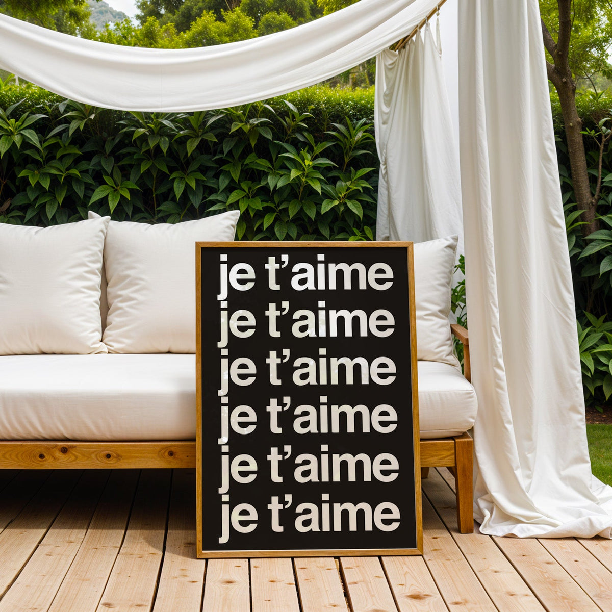 Je t'aime repeated six times in white type print in an oak frame on an outdoor daybed under white drapes