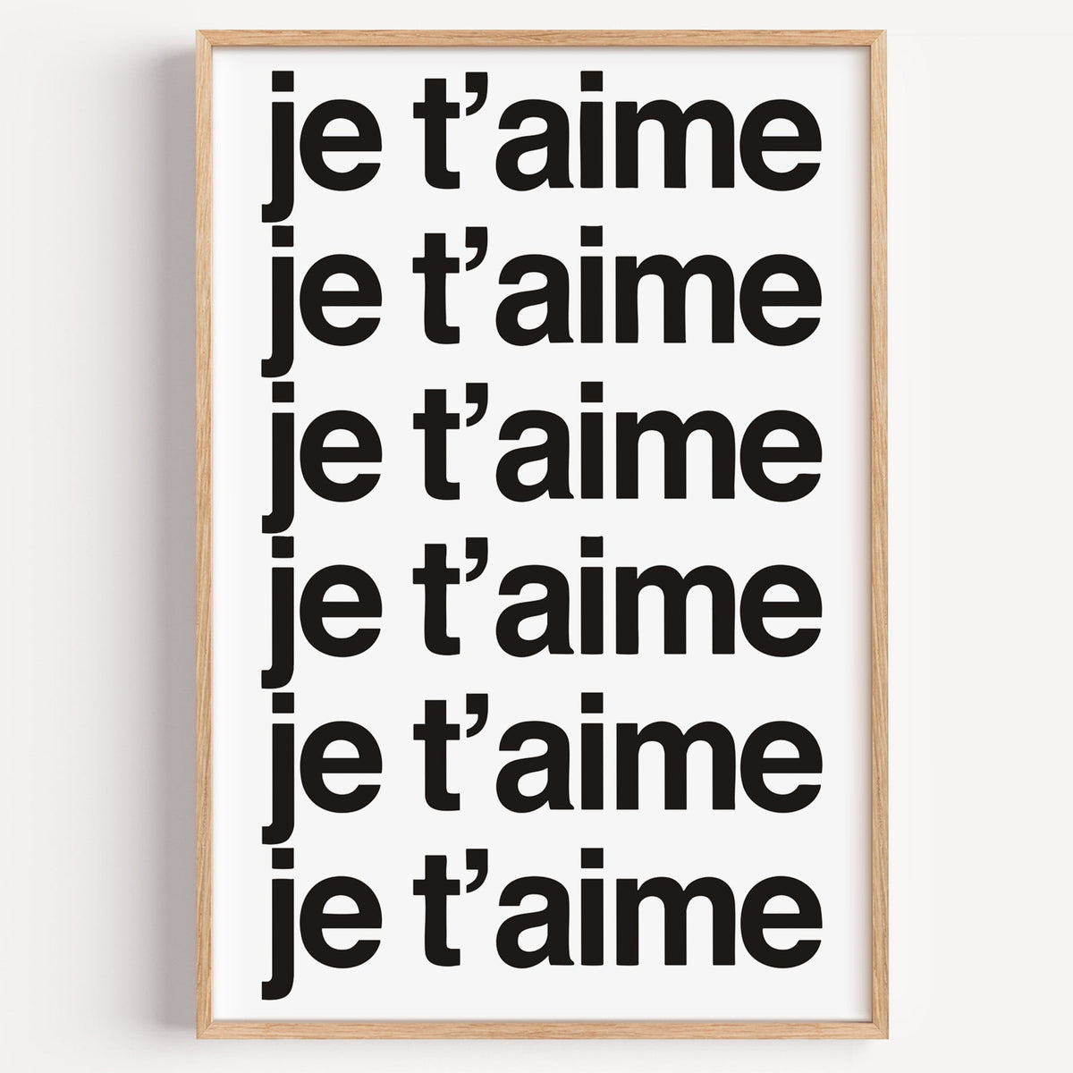 Je t'aime repeated six times in black type print in a light oak frame on a plain white wall