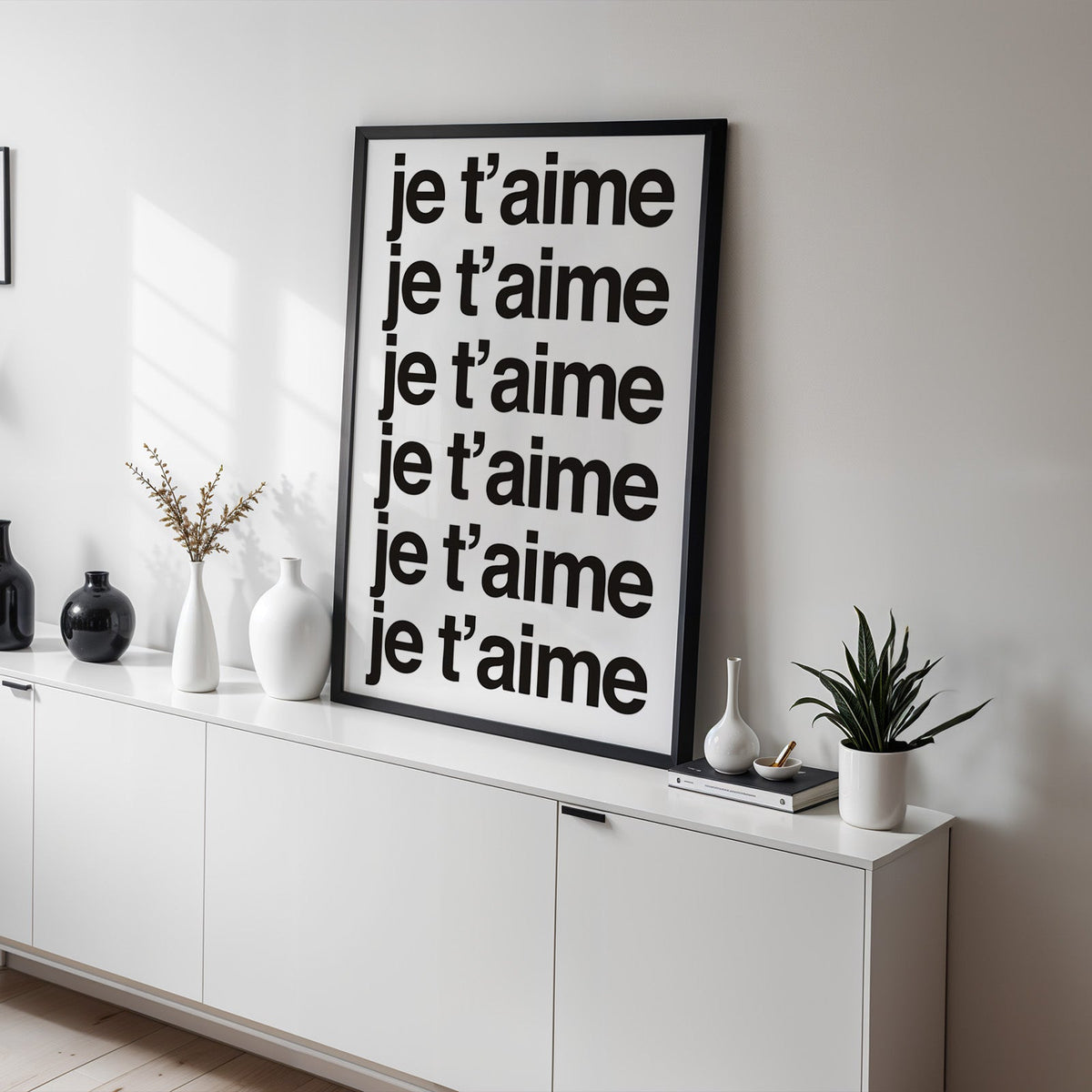 Je t'aime repeated six times in black type print in a black frame on a white sideboard with black and white vases