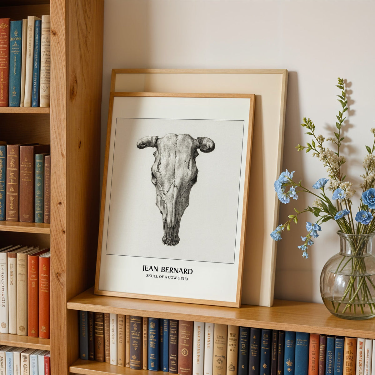 Cow skull print in an oak frame leaning on a bookshelf beside blue flowers