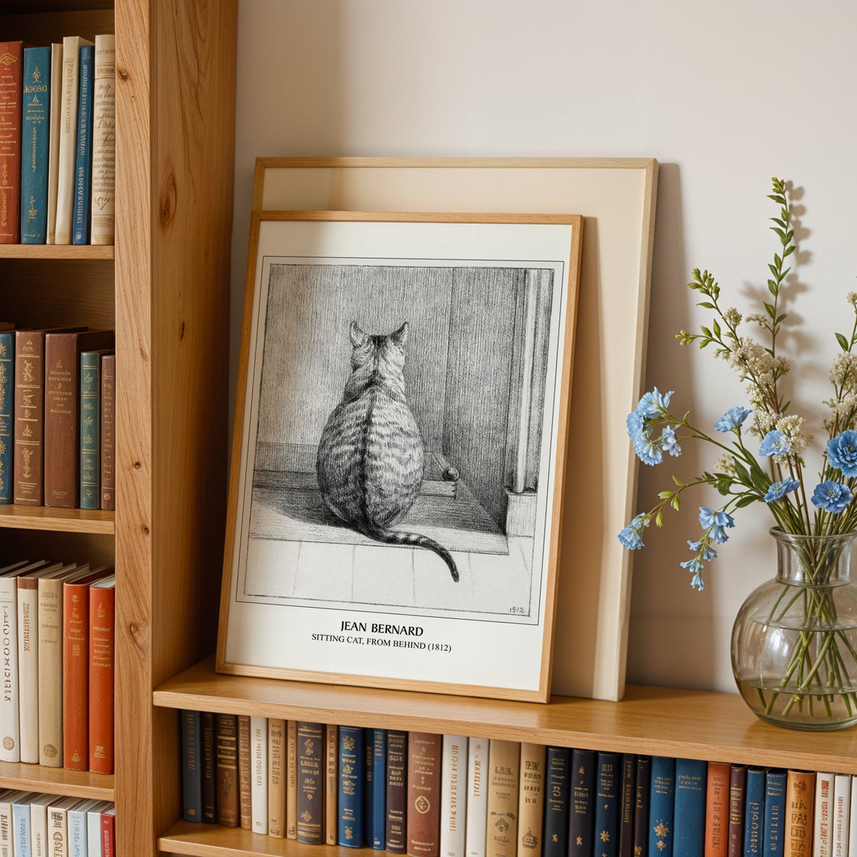 Sitting cat print in an oak frame leaning on a bookshelf beside blue flowers