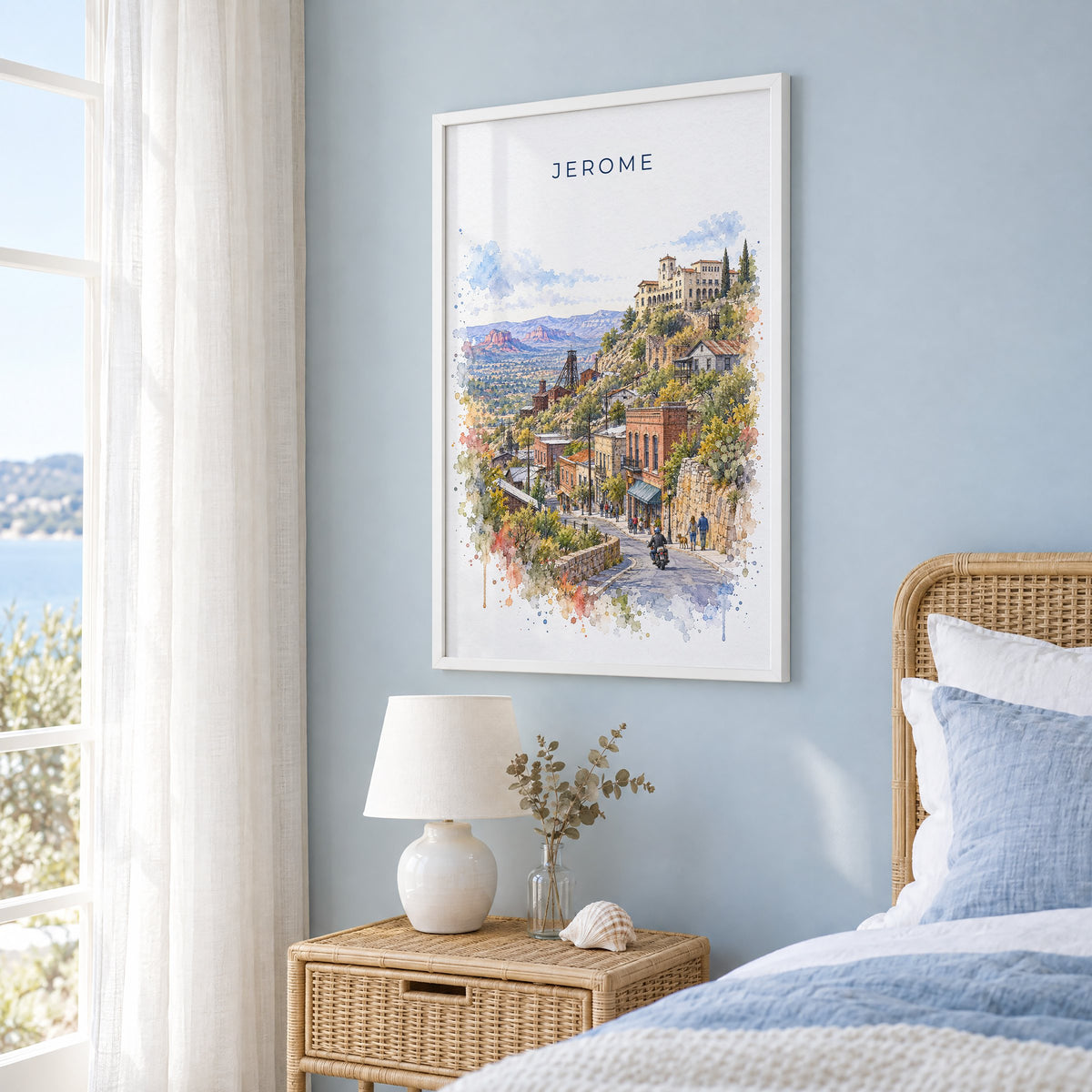 Jerome, Arizona watercolor print in a white frame on a pale blue wall beside a rattan bed in a coastal bedroom