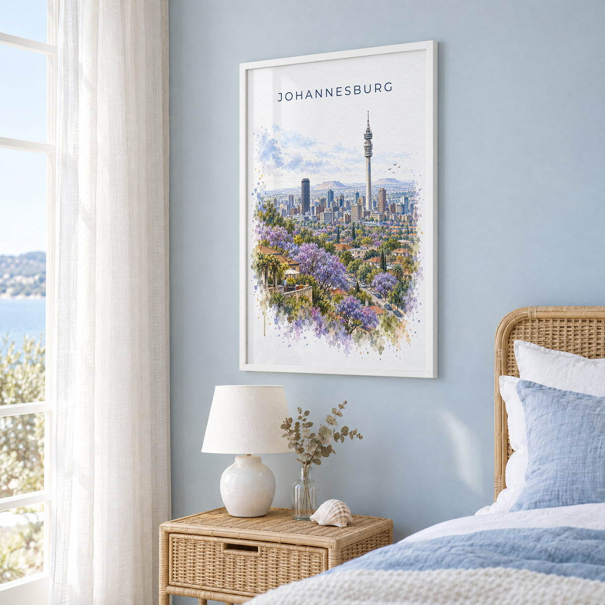 Johannesburg watercolor print in a white frame on a pale blue wall beside a rattan bed in a coastal bedroom