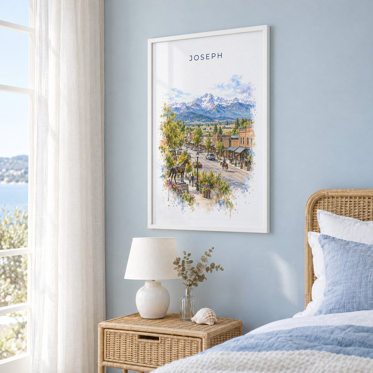 Joseph, Oregon watercolor print in a white frame on a pale blue wall beside a rattan bed in a coastal bedroom
