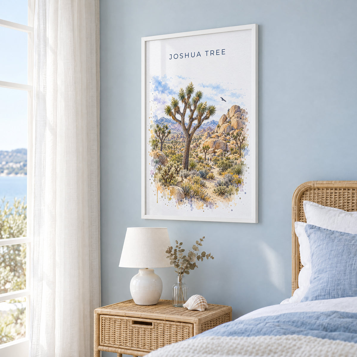 Joshua Tree National Park watercolor print in a white frame on a pale blue wall beside a rattan bed in a coastal bedroom
