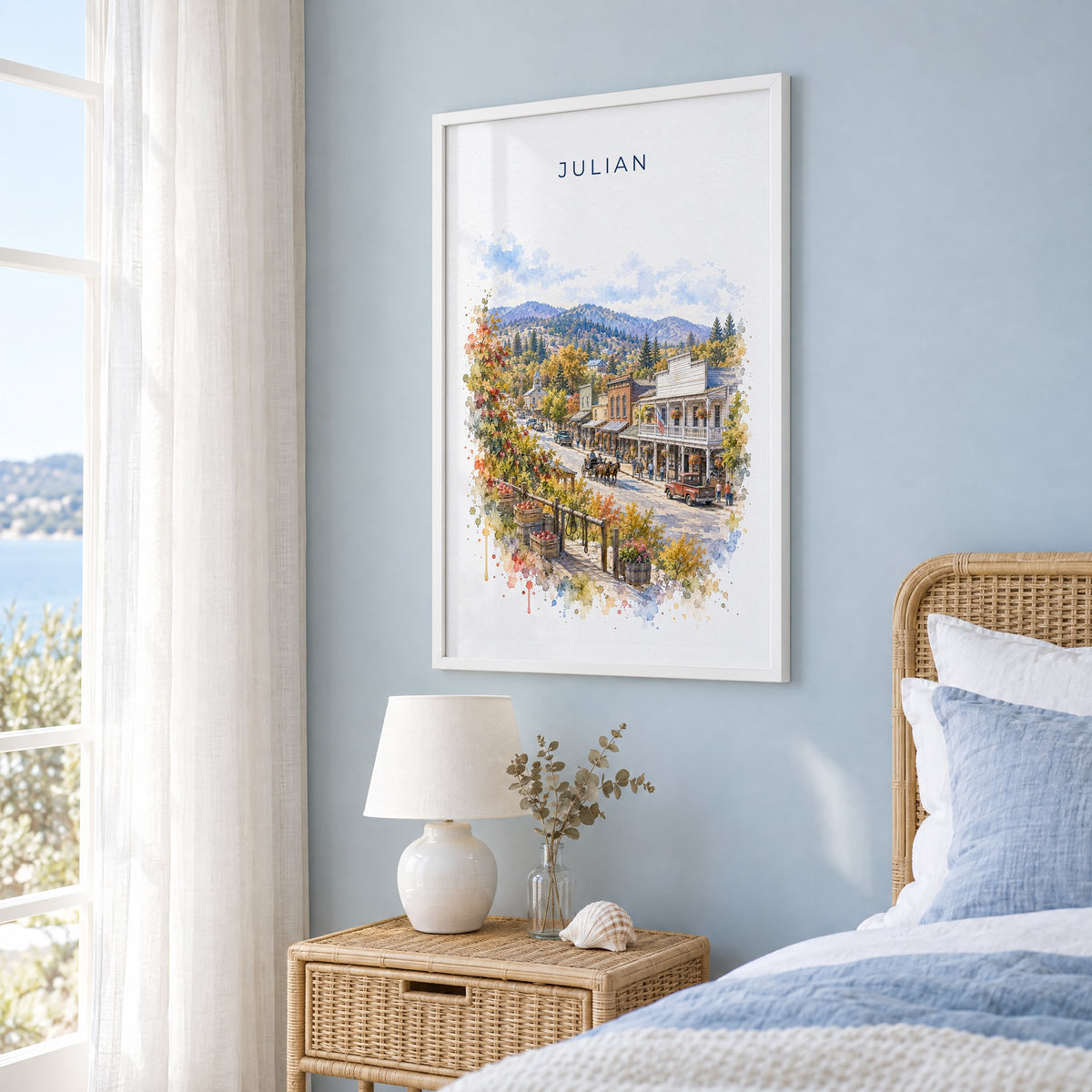 Julian, California watercolor print in a white frame on a pale blue wall beside a rattan bed in a coastal bedroom