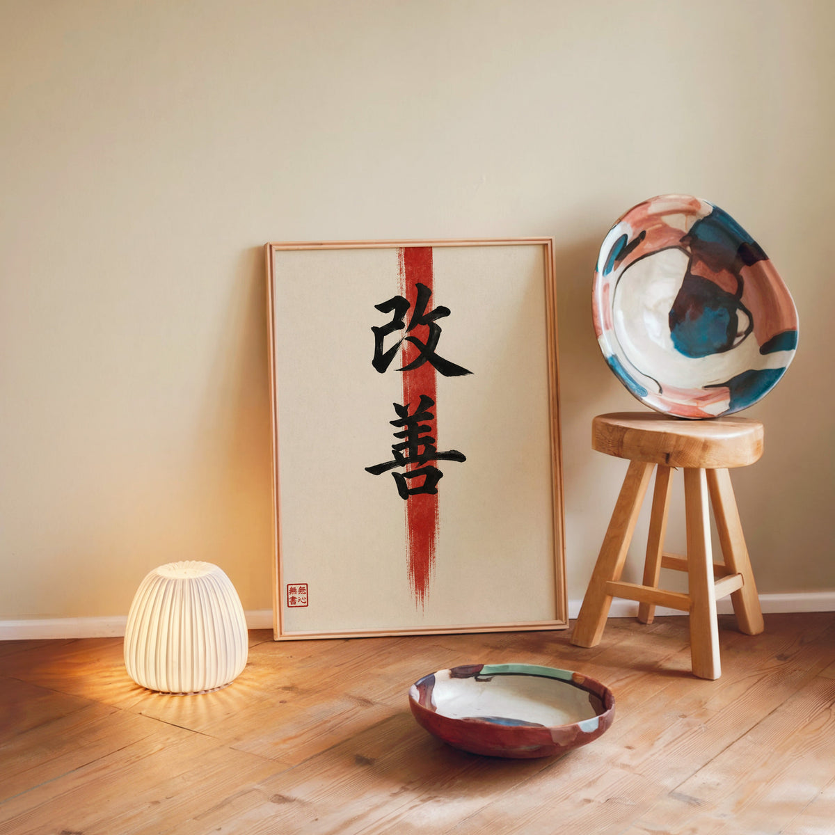Kaizen kanji calligraphy print in a light wood frame leaning on a wooden floor by a glowing lamp and stool