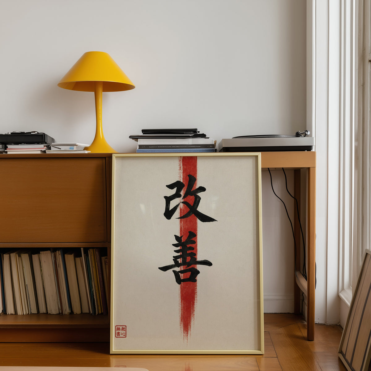 Kaizen kanji calligraphy print shown in a gold frame leaning under a teak desk with a yellow lamp