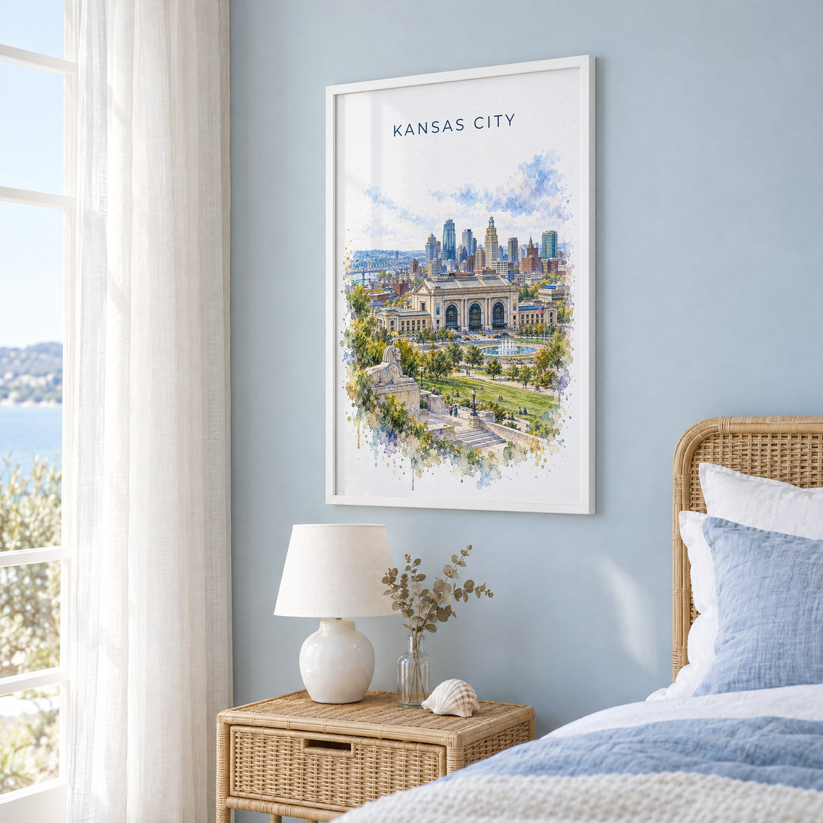 Kansas City watercolor print in a white frame on a pale blue wall beside a rattan bed in a coastal bedroom