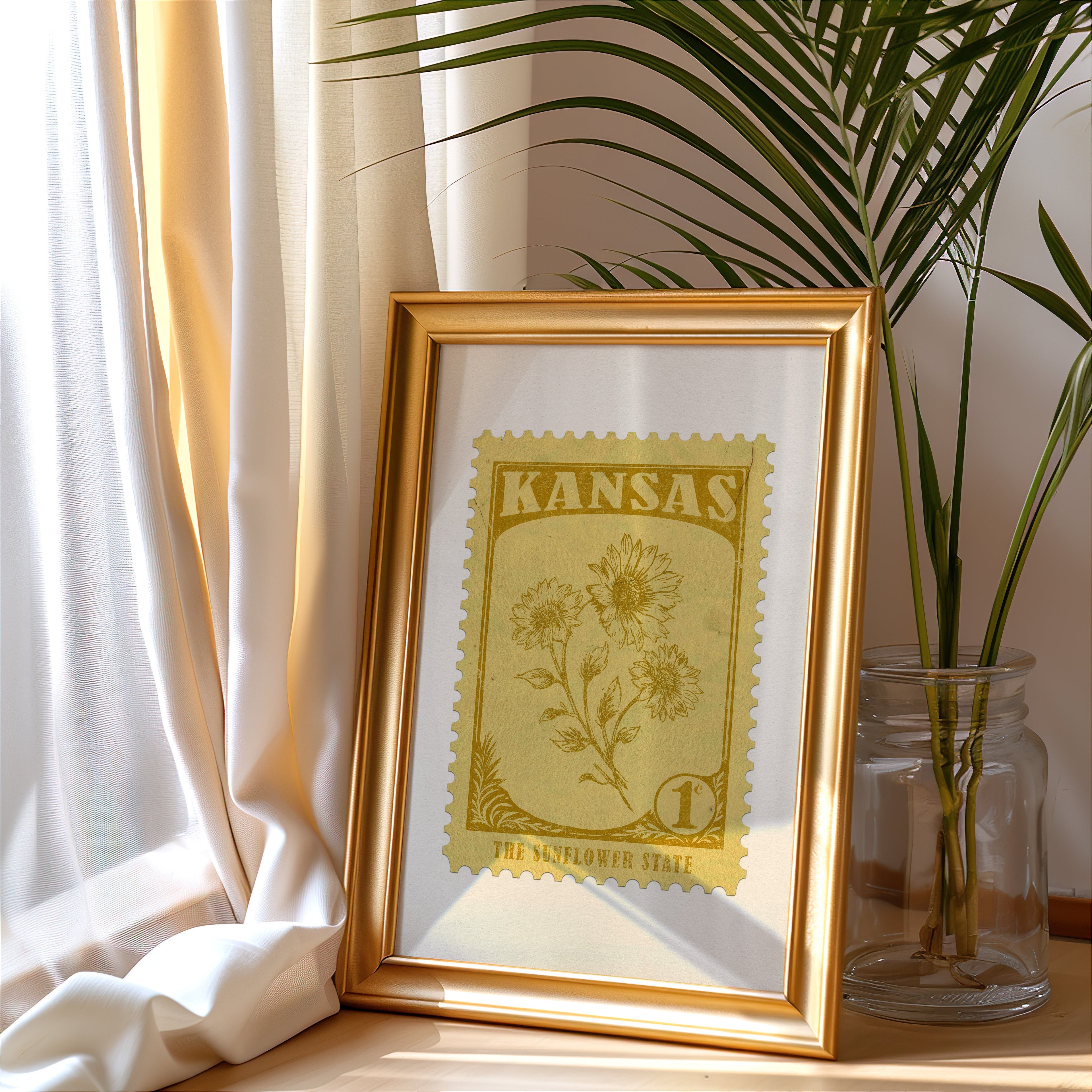 Kansas stamp print in a gold frame leaning by a cream curtain with a palm in a glass jar