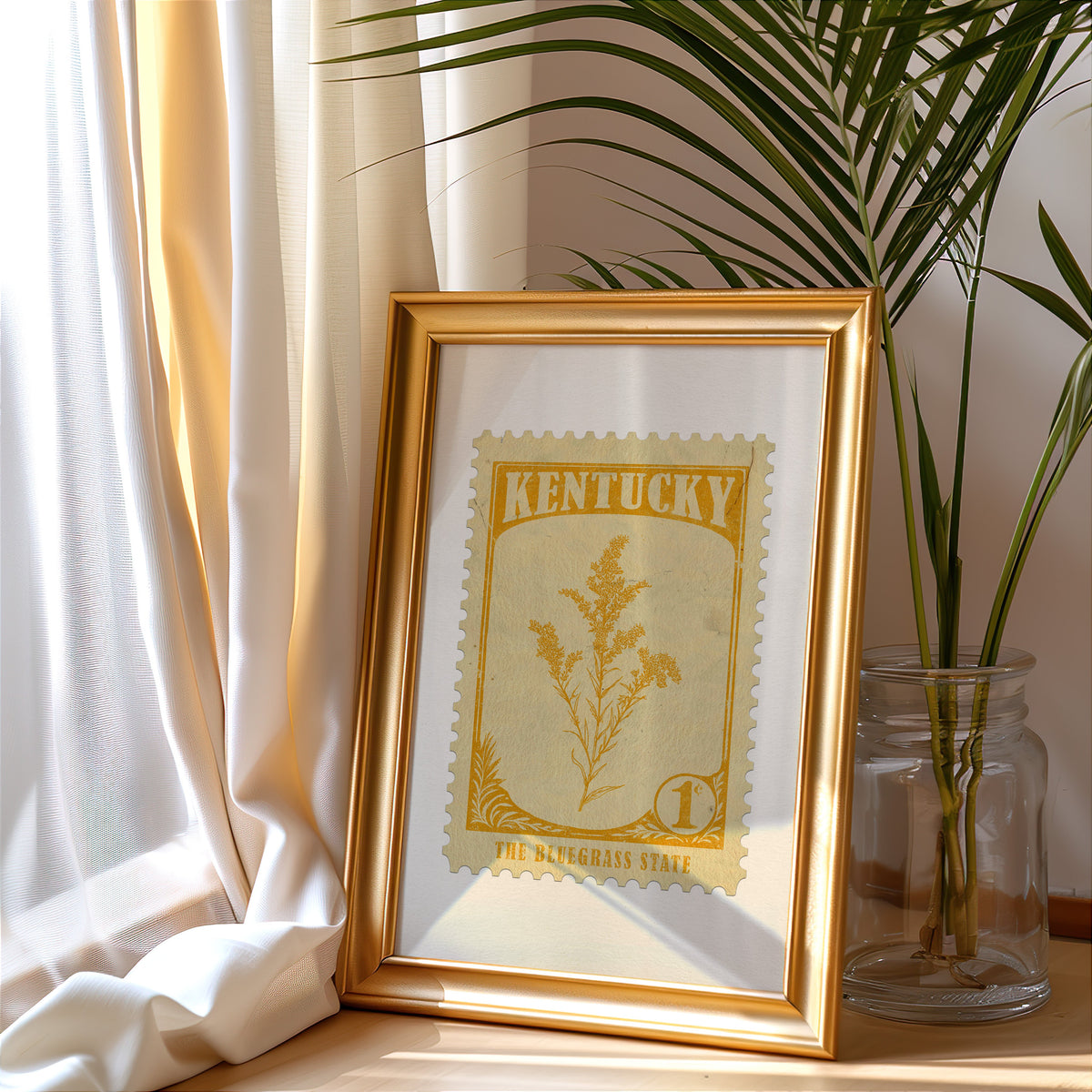 Kentucky stamp print in a gold frame leaning by a cream curtain with a palm in a glass jar