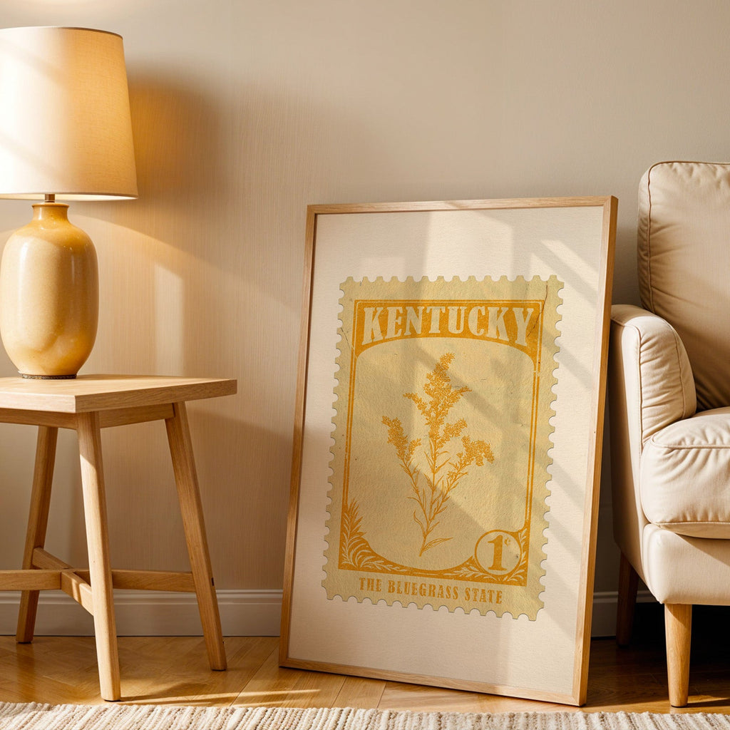 Kentucky stamp print in a wood frame resting on the floor beside a beige armchair and a yellow lamp