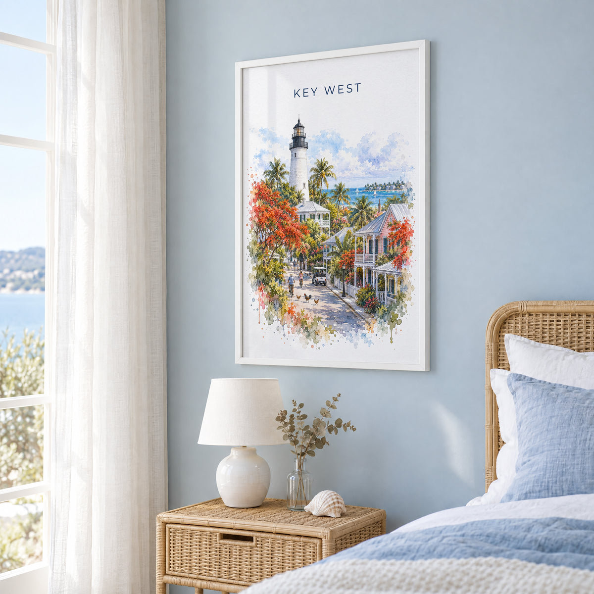 Key West watercolor print in a white frame on a pale blue wall beside a rattan bed in a coastal bedroom