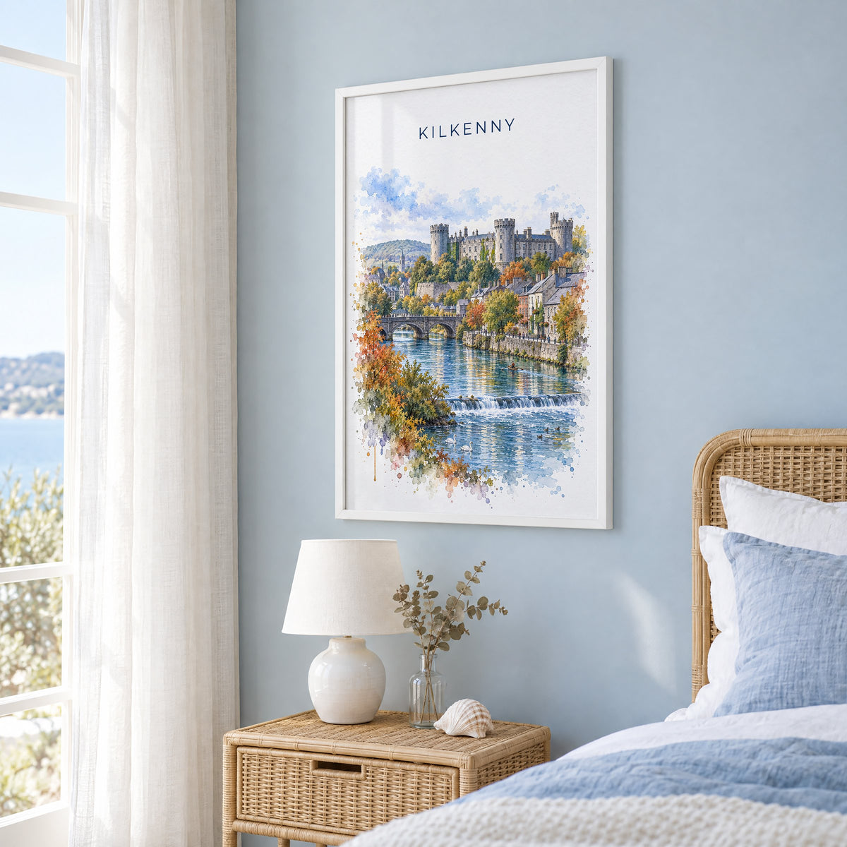 Kilkenny watercolor print in a white frame on a pale blue wall beside a rattan bed in a coastal bedroom