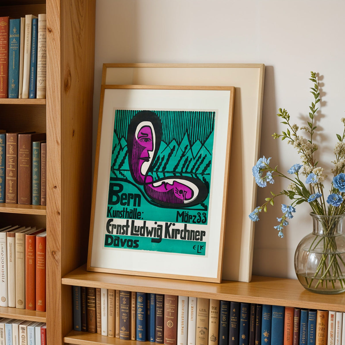 Exhibition poster print in an oak frame leaning on a bookshelf beside flowers