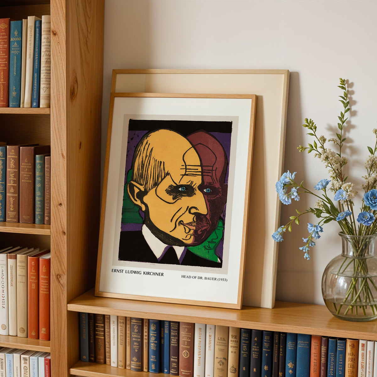 Expressionist head print in an oak frame leaning on a bookshelf beside flowers