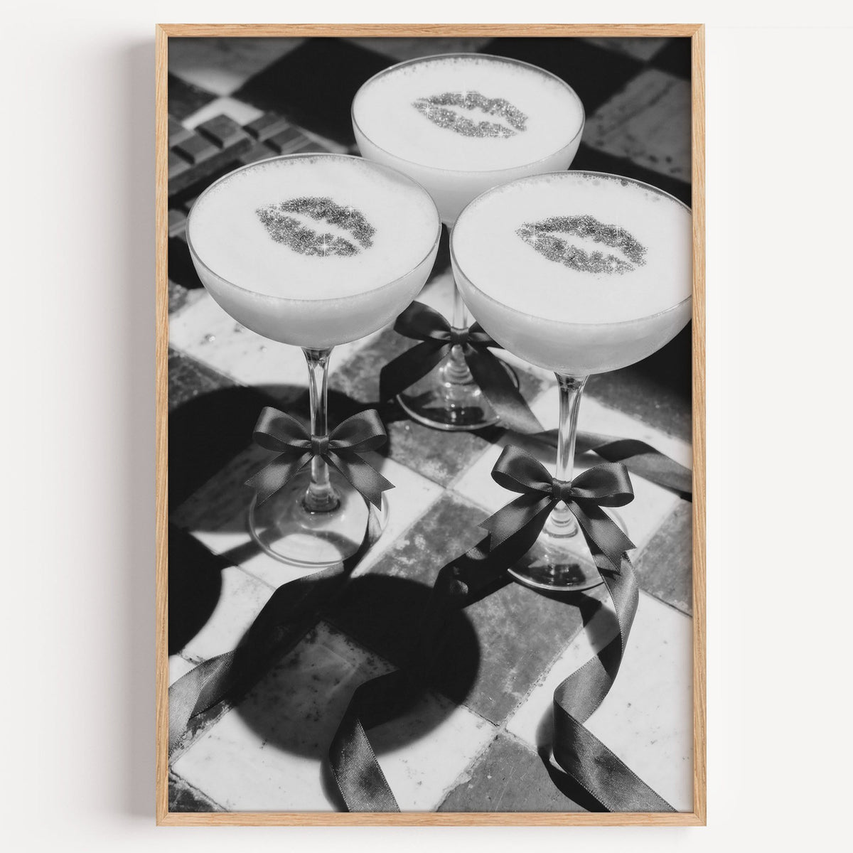 Three coupe cocktails with kiss marks and bows print in a light oak frame on a plain white wall