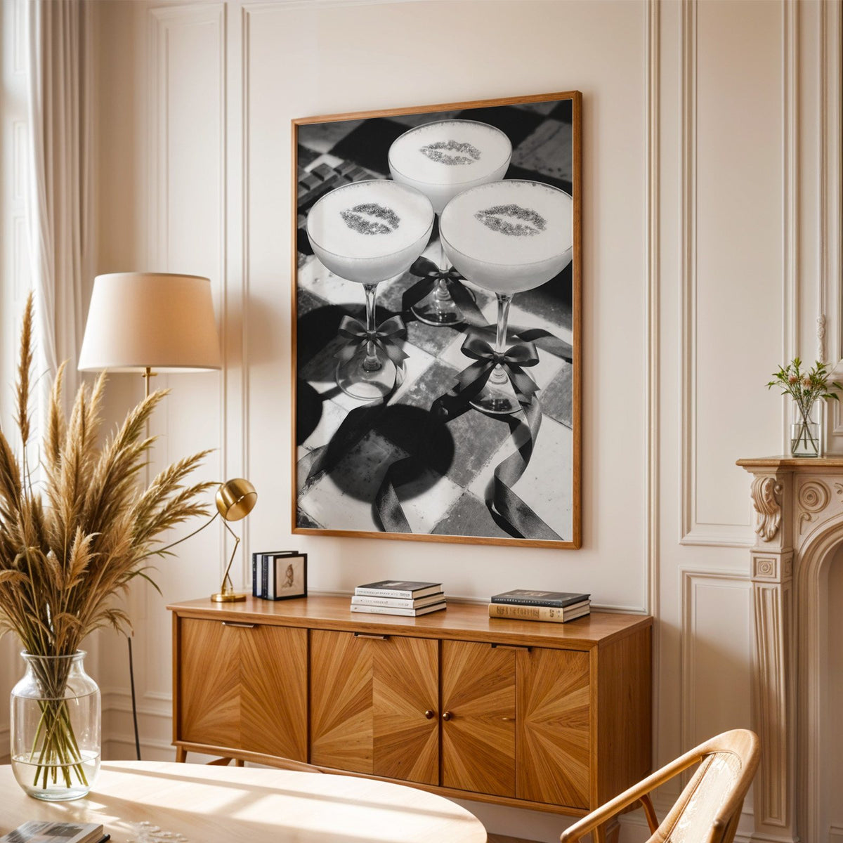 Three coupe cocktails with kiss marks and bows print in an oak frame above a sunburst wood sideboard with pampas grass