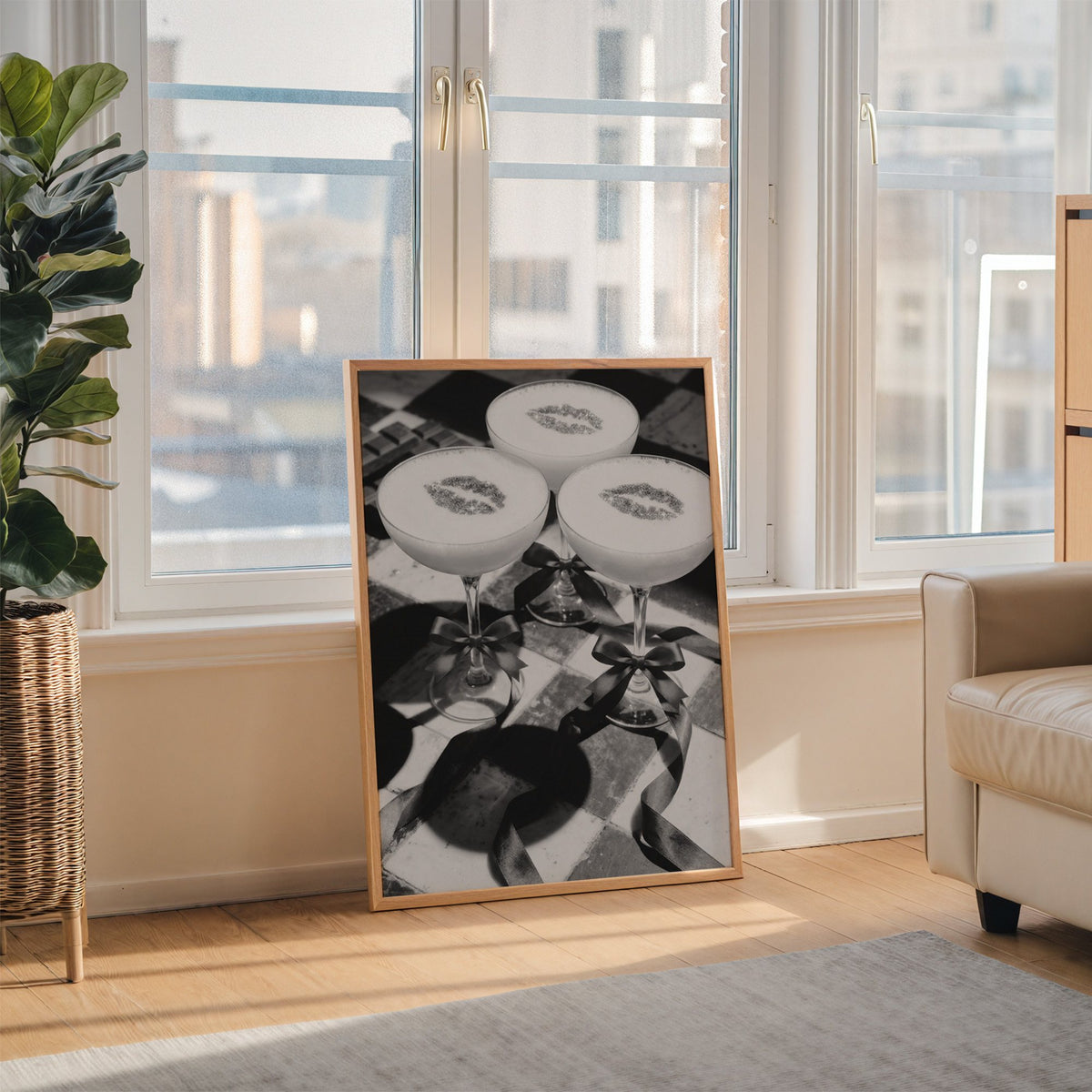Three coupe cocktails with kiss marks and bows print in an oak frame leaning on the floor beside a fiddle leaf fig