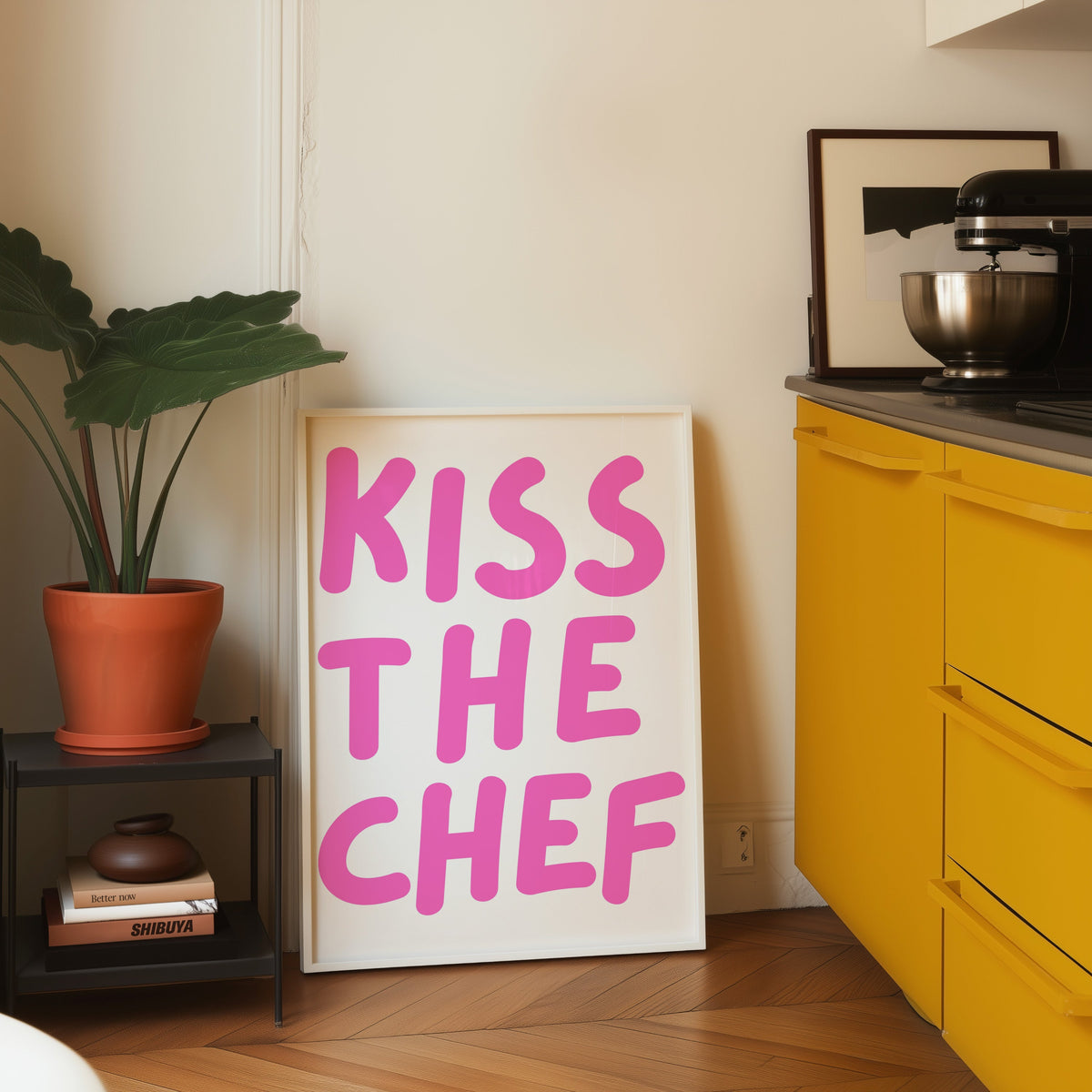 Kiss the Chef print in a white frame leaning on parquet beside a yellow kitchen
