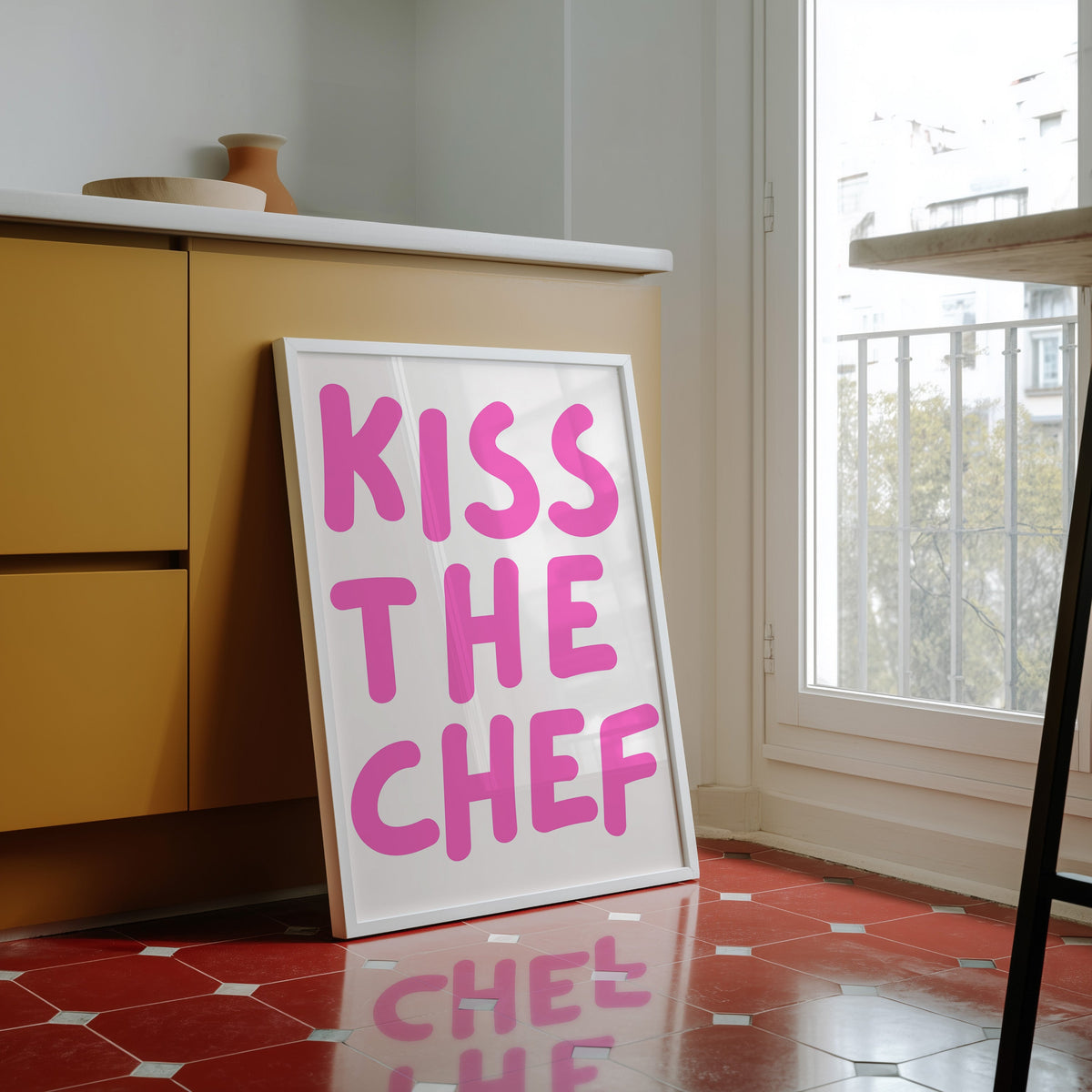 Kiss the Chef print in a white frame against yellow cabinets on a red tiled floor