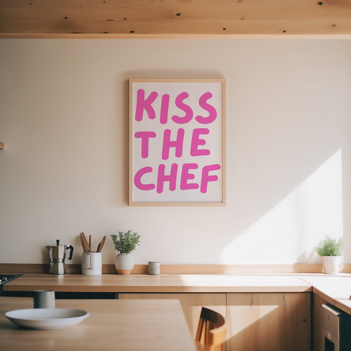 Kiss the Chef print in an oak frame above a wood counter with a moka pot and herbs
