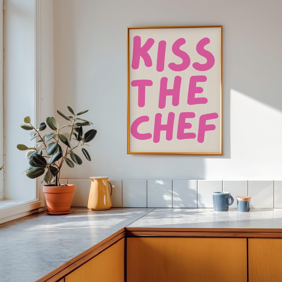 Kiss the Chef print in a gold frame above a marble counter with a yellow ceramic jug