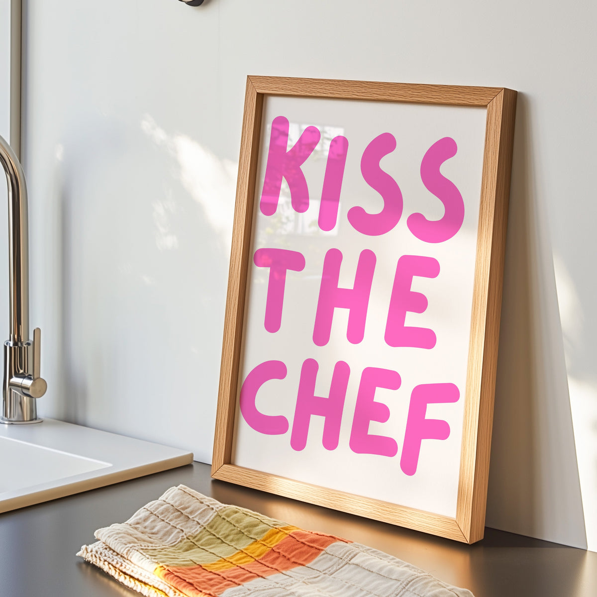 Kiss the Chef print in an oak frame by the kitchen sink beside a striped tea towel