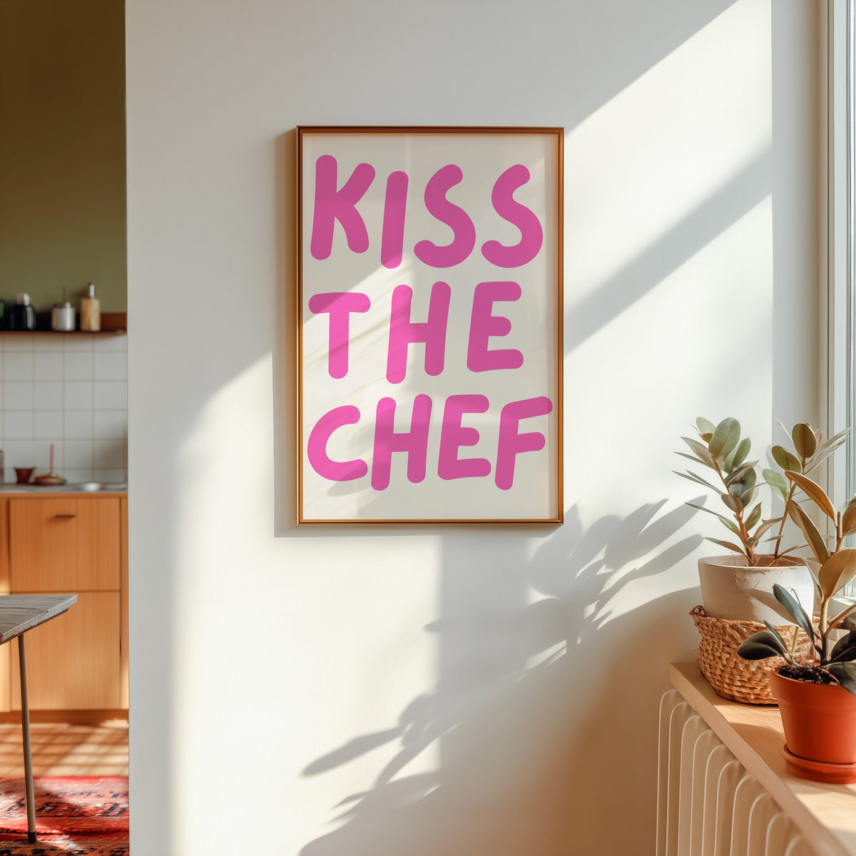 Kiss the Chef print in a gold frame in sunlight with plants and a kitchen beyond