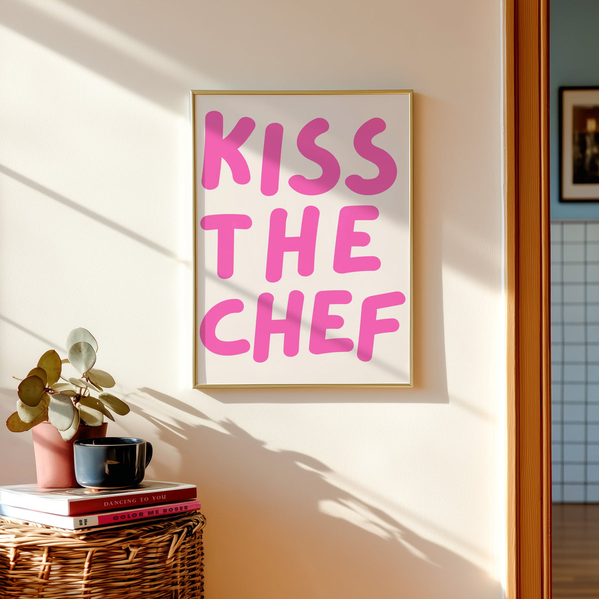 Kiss the Chef print in a gold frame above a basket of books and a pilea plant
