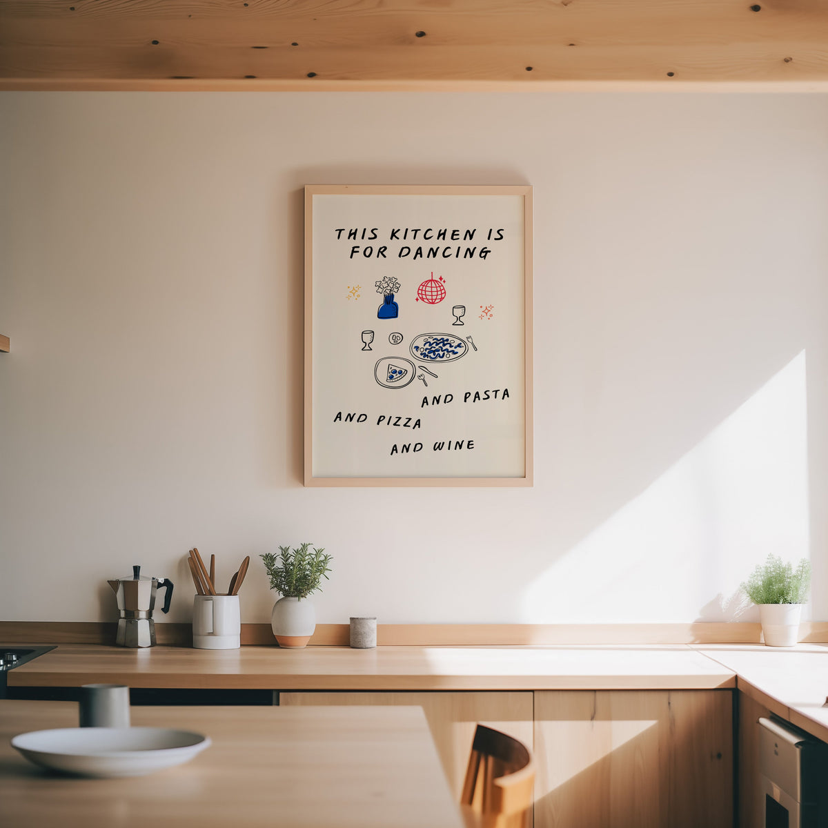 Kitchen Is For Dancing print in an oak frame above a wood counter with a moka pot and herbs
