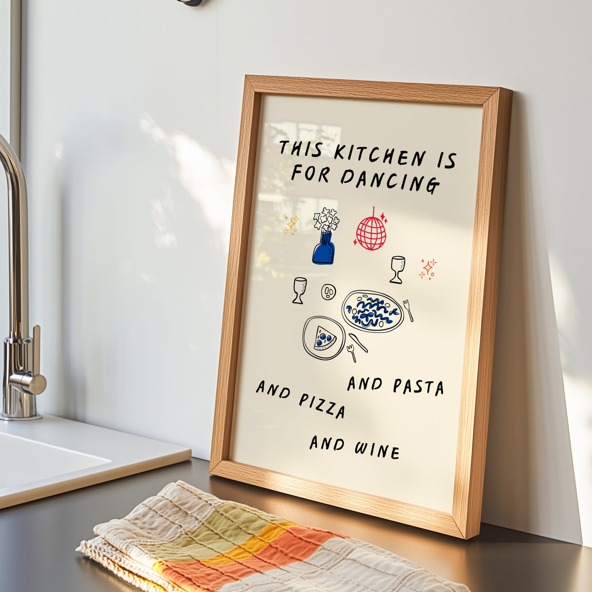 Kitchen Is For Dancing print in an oak frame by the kitchen sink beside a striped tea towel