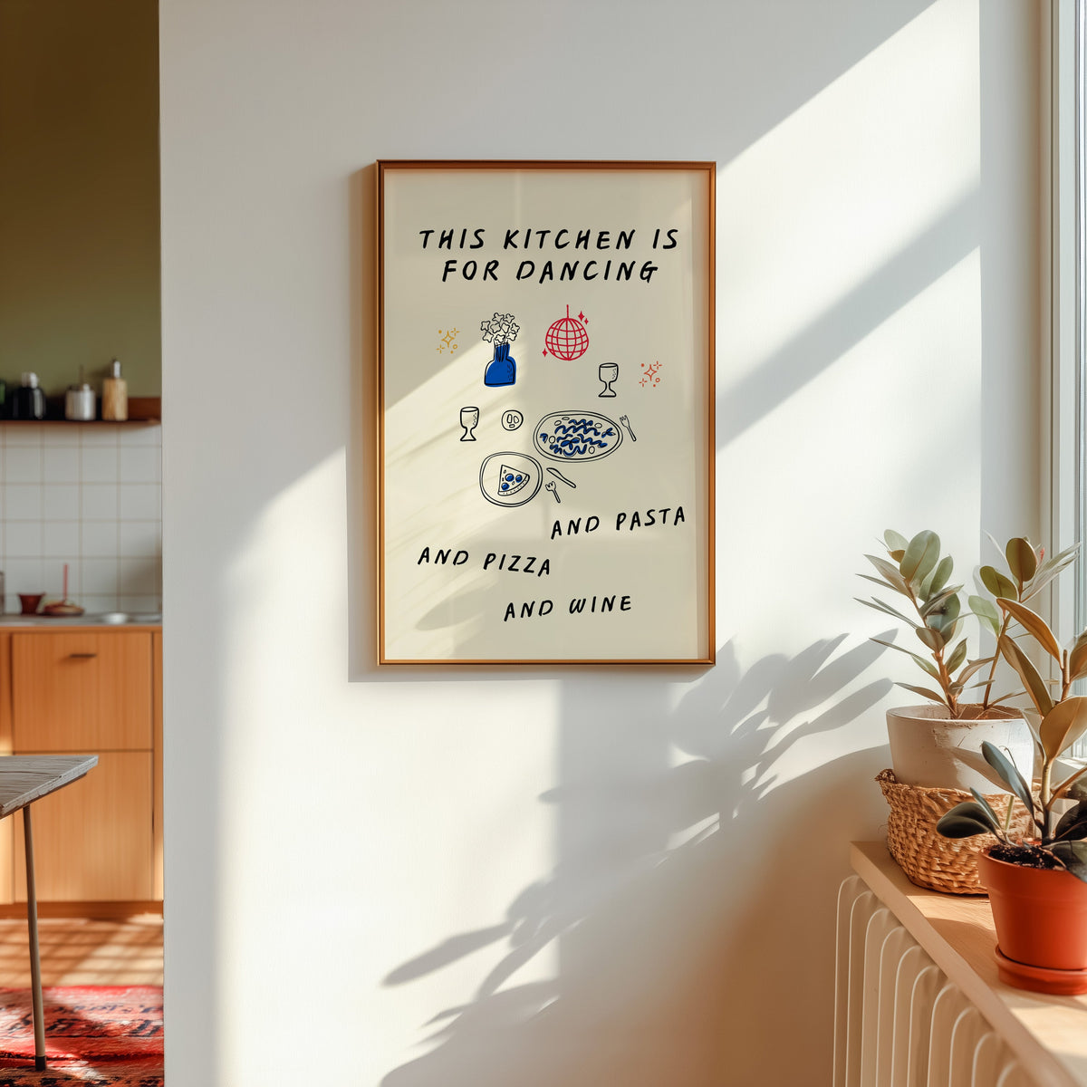 Kitchen Is For Dancing print shown in a gold frame in sunlight with plants and a kitchen beyond