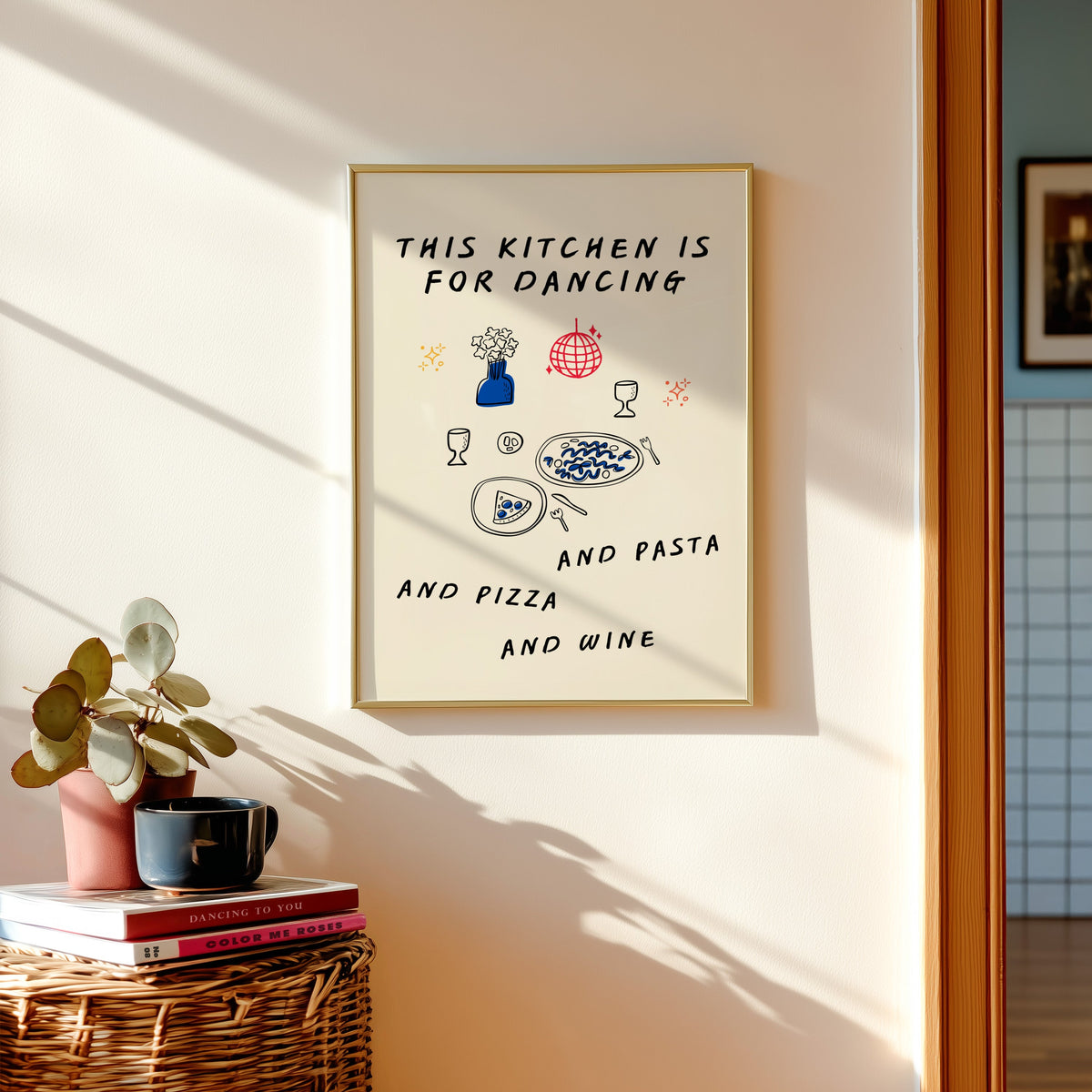 Kitchen Is For Dancing print shown in a gold frame above a basket of books and a pilea plant