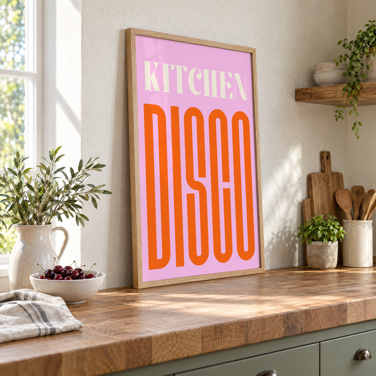 Kitchen Disco pink and orange type print in an oak frame leaning on a butcher block island beside a bowl of cherries
