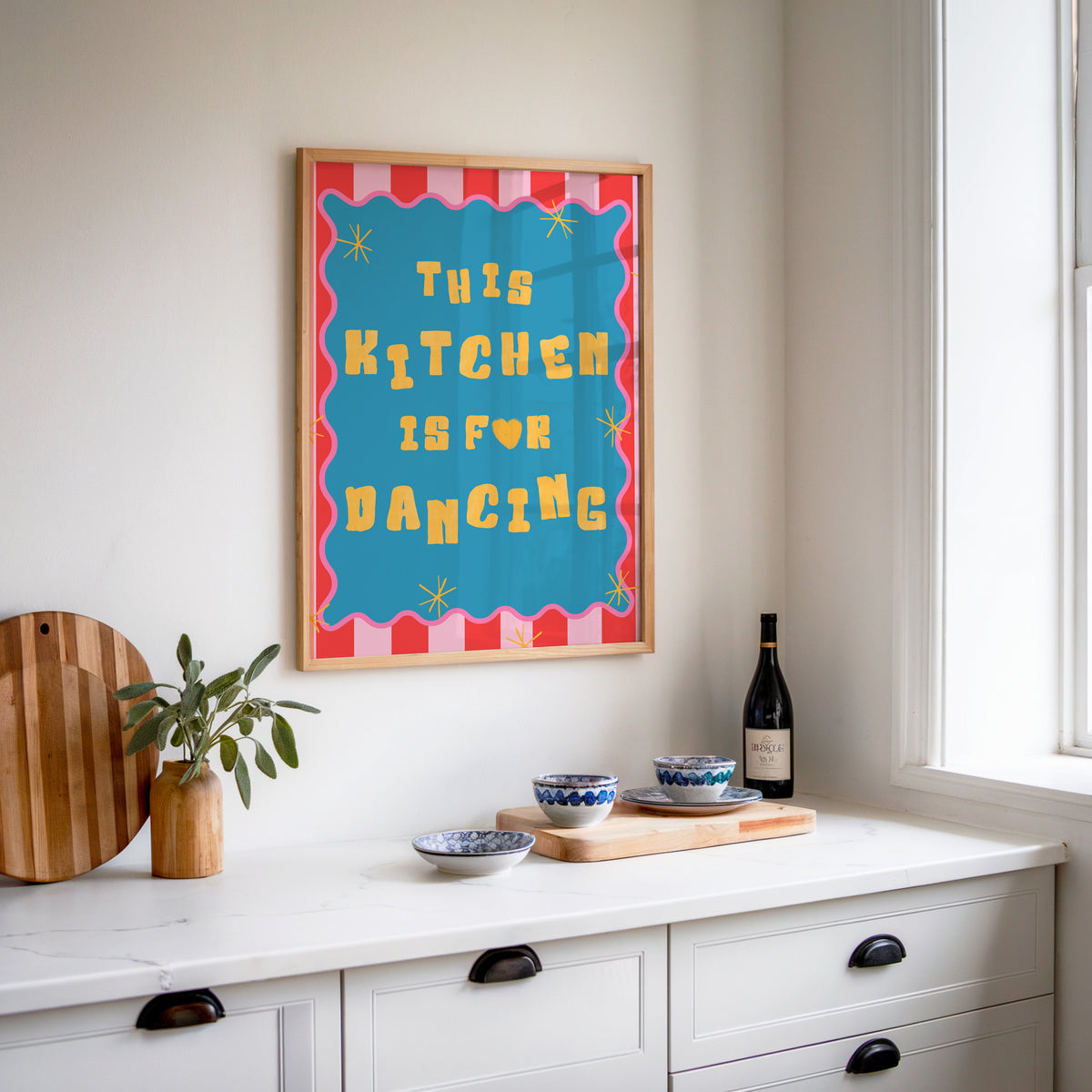 This Kitchen Is For Dancing print in an oak frame above a white sideboard with a bottle of wine